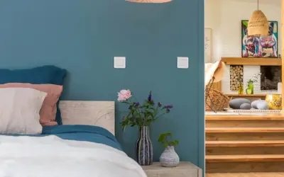 Un lit douillet avec des coussins roses et blancs sur un mur couleur bleu canard. À côté, un escalier en bois mène à un salon cosy.