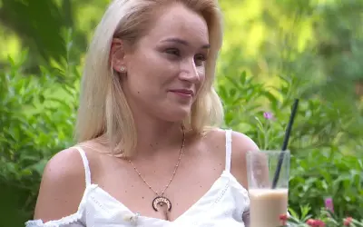 Uśmiechnięta Kamila w białej sukience, z naszyjnikiem z księżycem, popija kawę mrożoną w letnim ogrodzie.