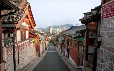 En smal gata kantad av traditionella hus med tegeltak leder mot en modern stadssilhuett i Seoul, Sydkorea.