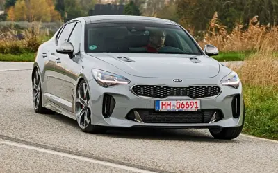 Szary Kia Stinger z kierowcą za kierownicą jedzie po krętej drodze, otoczony jesienną roślinnością.