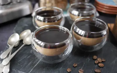 Fyra små glas med kaffe pannacotta, toppad med mörk sirap. Tre skedar ligger till vänster, och kaffebönor sprider sig på stenen.
