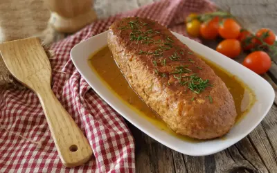Polpettone in padella dorato e succulento, guarnito con erba cipollina, pronto per essere servito. Accanto, una spatola in legno e pomodorini freschi.