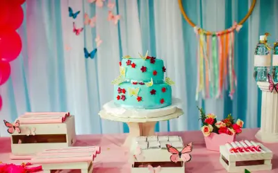 Torta a due piani decorata con farfalle e fiori in pasta di zucchero, su un tavolo rosa con palloncini rossi e caramelle.