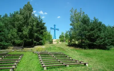 Niebieski krzyż na wzgórzu, najwyższy szczyt wielkopolski, otoczony drzewami. Poniżej amfiteatr z drewnianymi ławami.