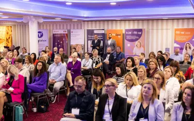 Sala konferencyjna pełna ludzi siedzących w rzędach, z banerami sponsorów w tle.