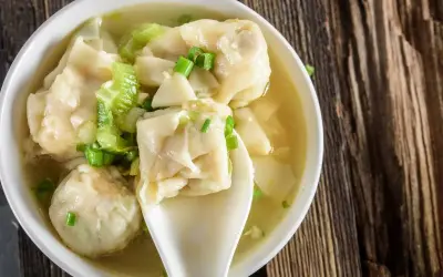 Aromatyczna zupa wonton z zieloną cebulką i kawałkami selera, podana w białej miseczce na drewnianym blacie.