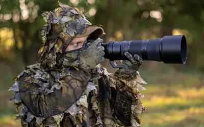 Fotograf w ghillie suit z aparatem do fotografowania zwierząt czeka na idealny kadr.
