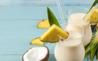 Dwa orzeźwiające koktajle Pina Colada, ozdobione kawałkami ananasa i liśćmi, na tle niebieskich desek. Idealny przepis na lato!