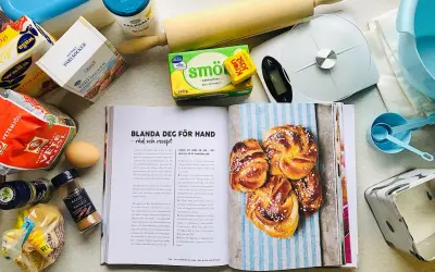 Här finns allt för ett lyckat kanelbullar recept: mjöl, jäst, smör, socker, kardemumma och kanel. En öppen bok visar hur man gör.