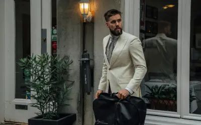 Mann im cremefarbenen Anzug mit gemustertem Hemd und Krawatte, trägt eine schwarze Kleidersacktasche. Ein lässiges Outfit für unterwegs.