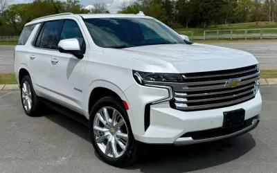 Biały Chevrolet Tahoe z chromowanymi felgami stoi na drodze, otoczony zielenią i chmurami.