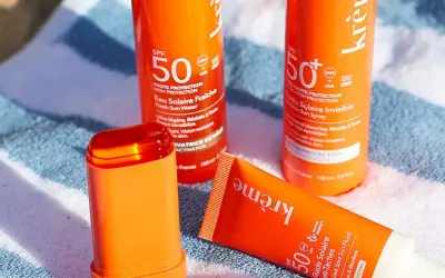 Trois produits solaires orange vif, dont un stick, sur une serviette rayée. Idéal pour savoir quelle crème solaire choisir pour une haute protection SPF 50.