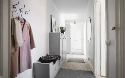 En smal hall med en rosa kappa och en vit halsduk på krokar. En grå förvaringsmöbel med en fårskinnspläd.