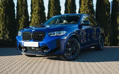 Błękitny suv BMW X4 M Competition błyszczy na tle zielonych drzew.
