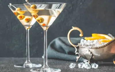 Dwa kieliszki martini z oliwkami, obok srebrna miseczka z lodem i cytryną. Idealne do podania z czym pić martini.