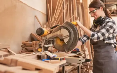 Artisane souriante utilisant une scie à onglet. Elle porte des lunettes et un tablier, des éléments essentiels pour comment protéger une création artisanale.