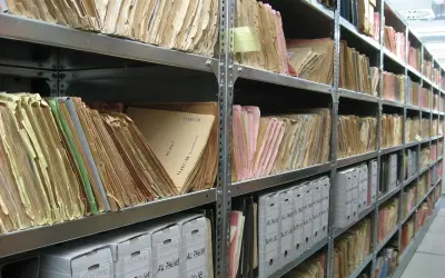 Rzędy półek archiwum wypełnione teczkami i pudłami. Na jednej z teczek widoczna jest sygnatura: "11497/58".