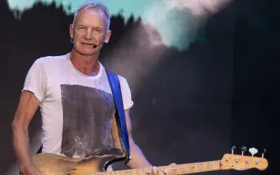 Sting gra na gitarze basowej w białej koszulce na tle sceny.