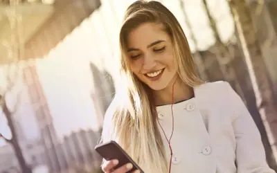 Uśmiechnięta blondynka w białym płaszczu słucha muzyki na telefonie. To inspiracja, jak być kobiecą.
