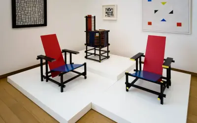 Twee iconische stoelen in rood, blauw en zwart, kenmerkend voor bauhaus kunst, staan naast een abstract schilderij en een sculptuur.