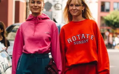 Dwie kobiety w sportowych stylizacjach, idealnych na podróż. Różowa bluza i granatowe dresy lub pomarańczowa bluza i czerwone dresy to świetny pomysł, jak ubrać się do samolotu.