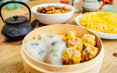 Pyszne dim sum w bambusowym parowniku, obok miseczki z makaronem i daniem mięsnym.