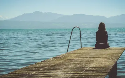 Mujer sentada en un muelle, contemplando el lago y las montañas. Pensamientos de frases de preocuparse por alguien flotan en el aire.