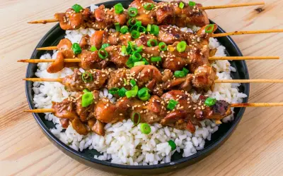 Fyra kycklingspett teriyaki ligger på en bädd av vitt ris, toppade med sesamfrön och hackad salladslök.