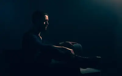 Mężczyzna z brodą siedzi w półmroku, wpatrzony w dal. Jego postawa jest **low key**, emanuje spokojem i zadumą.