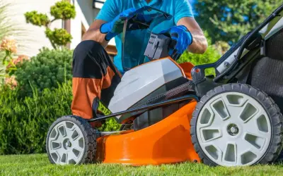 Mężczyzna wymienia baterię w pomarańczowej kosiarce akumulatorowej, przygotowując się do pracy w ogrodzie. To ważny element w rankingu kosiarek akumulatorowych.