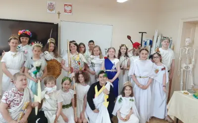 Dzieci w strojach antycznych bogów i bogiń, jak Artemida, pozują do zdjęcia. W tle szkielet i tablica.