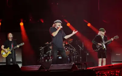 Zespół AC/DC na scenie, wokalista śpiewa, gitarzyści grają. Czerwone światła.