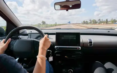Vue du **rétro intérieur position nuit** d'une voiture. Mains sur le volant, route dégagée sous un ciel nuageux.