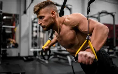 Mężczyzna wykonuje pompki z użyciem taśm TRX. Intensywny trening okluzyjny buduje siłę i masę mięśniową.