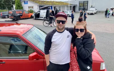 Ett par poserar framför en röd bil, redo att bygga ljud i bil. I bakgrunden syns bilar och folk vid en butik.