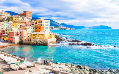Case colorate a picco sul mare turchese, barche ormeggiate e un promontorio roccioso: un angolo di Genova che ricorda le sue spiagge più belle.