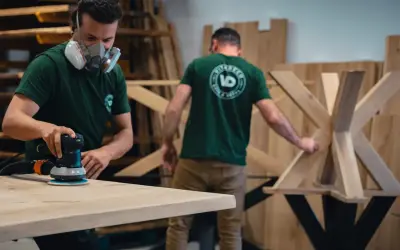 Un artisan masqué utilise une ponceuse pour poncer une table en bois. Un autre artisan travaille sur un pied de table en bois en arrière-plan.