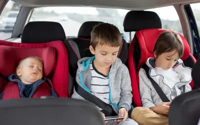 Dziecko śpi w foteliku, a dwaj starsi bracia grają na telefonach. Od kiedy można zapinać dziecko pasami samochodowymi? Od urodzenia, w odpowiednim foteliku.