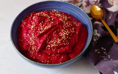 Hummus di barbabietola cremoso e vibrante, cosparso di semi di sesamo, in una ciotola blu. Un cucchiaio dorato e un tovagliolo fiorato completano la scena.