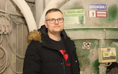 Mężczyzna w okularach i czarnej kurtce z futrzanym kapturem stoi na tle przemysłowych maszyn.
