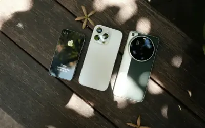 Trzy telefony do zdjęć: stary iPhone, nowy iPhone i smartfon z aparatem Leica, leżące na drewnianym stole.