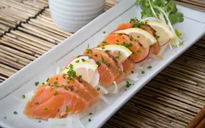 Tataki de salmón fresco, finamente cortado y adornado con rodajas de limón, rábano y cebollino, servido sobre una cama de finas hebras de cebolla.