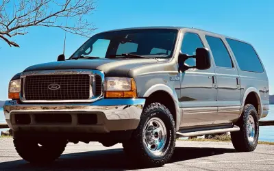 Brązowy Ford Excursion z chromowanymi dodatkami i terenowymi oponami stoi na tle błękitnego nieba i nagich drzew.