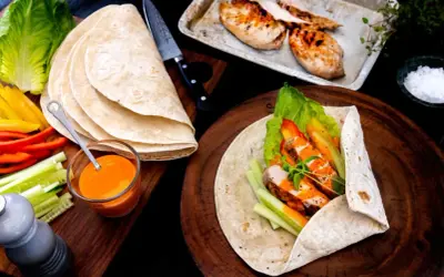 En färdig kyckling wrap med sallad, paprika och gurka. Bredvid ligger tortillabröd, dressing och grillad kyckling.