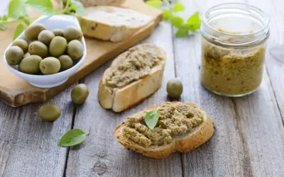 Bruschette con crema di olive, un vasetto di crema di olive e olive verdi su un tagliere di legno.
