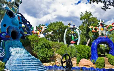 Sculture colorate e fantastiche popolano il **giardino dei tarocchi**, tra alberi verdi e cielo nuvoloso.