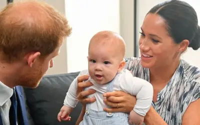 Meghan Markle trzyma swoje dziecko, które z zaciekawieniem patrzy na księcia Harry'ego.