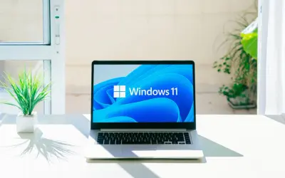 Laptop z logo Windows 11, gotowy na aktualizację 25H2. Na biurku obok rośliny, w jasnym pomieszczeniu.