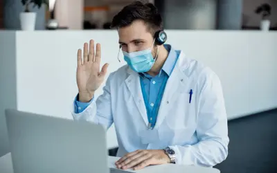 Lekarz w maseczce i kitlu wita pacjenta podczas konsultacji telemedycznej.