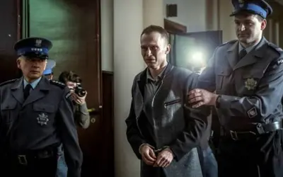 Scena z filmu o Tomaszu Komendzie: policjanci eskortują go w kajdankach, a za nimi stoi fotograf z aparatem.